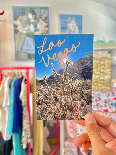 A scenic landscape photograph of Redrock Canyon featuring rocky formations and desert vegetation. The words ‘Las Vegas’ are artistically drawn on the image, blending natural beauty with urban charm.