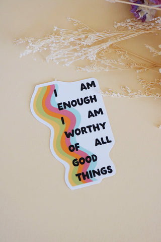 Sticker that reads “I am enough I am worthy of all good things”. The letters are layered over a rainbow colored squiggle graphic